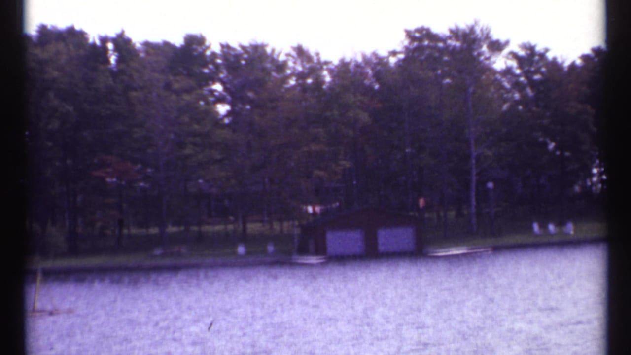 Lake water facing a wooded shore with a small boathouse and lawn chairs.