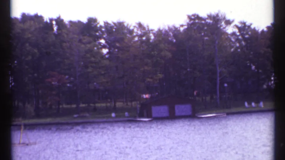 1967 8mm frame of a quiet Montreal waterfront with water in front, trees behind, and a small boathouse near the bank.