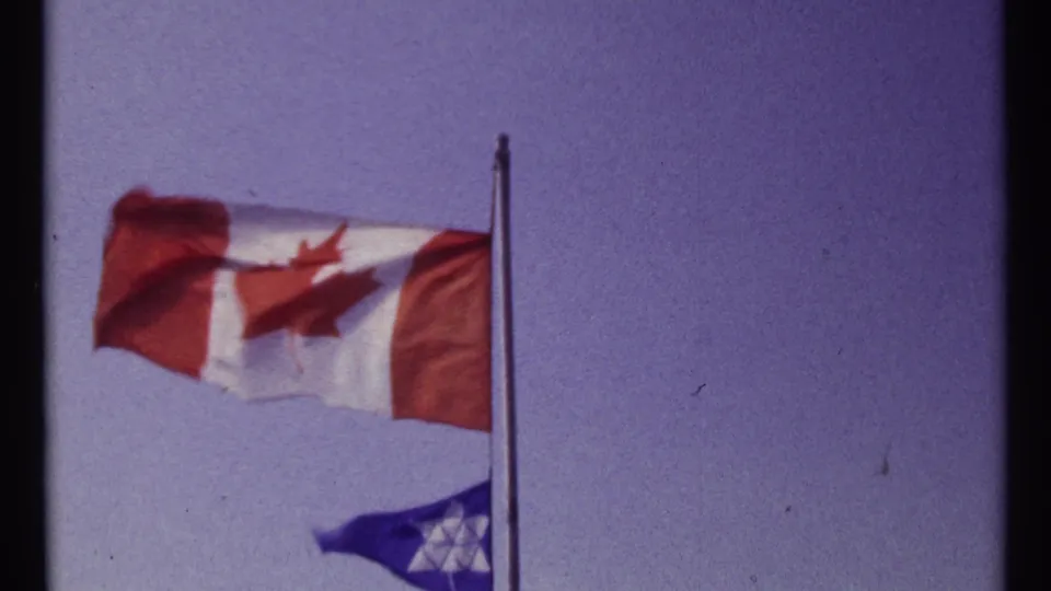 A close 8mm view of the Canadian flag with a smaller blue Star of David flag below, both blown sideways by wind.