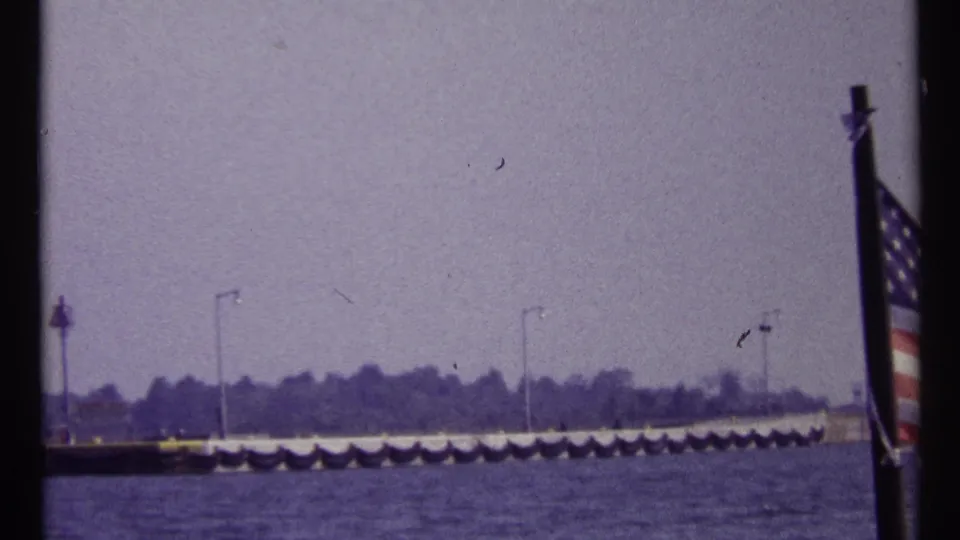 Montreal-linked waterfront frame with water, a long pier or breakwater, distant trees, and a partial striped flag at right.
