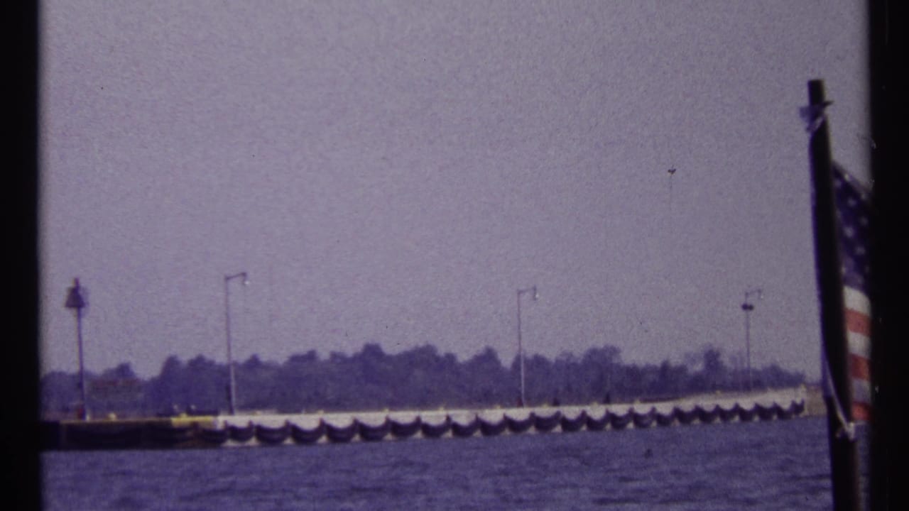 1967 Montreal harbor view with an American flag in the foreground, water below, and a distant pier with lampposts under a hazy sky.