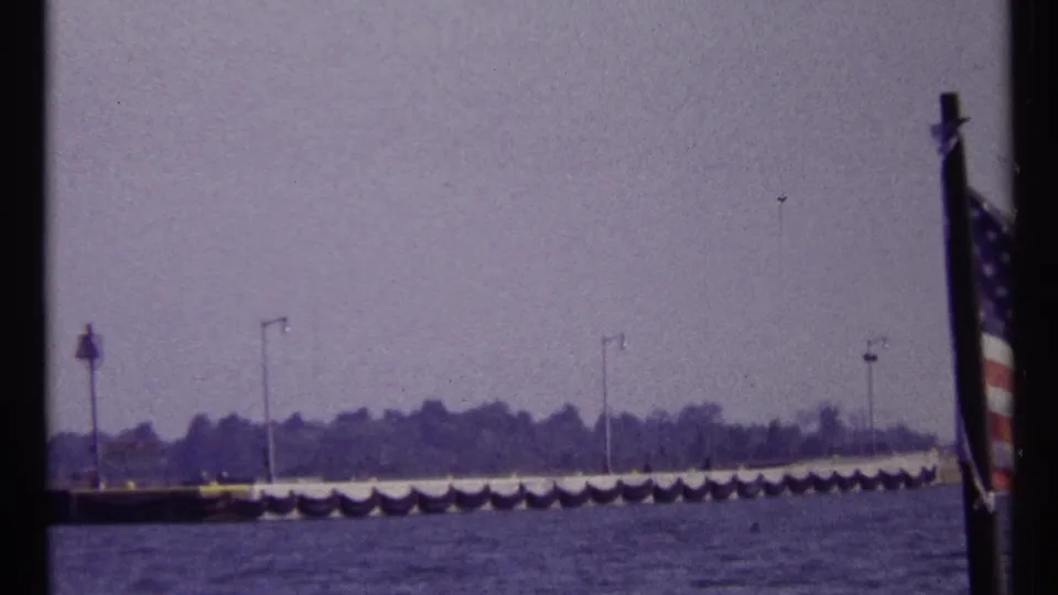 1967 Montreal harbor view with an American flag in the foreground, water below, and a distant pier with lampposts under a hazy sky.