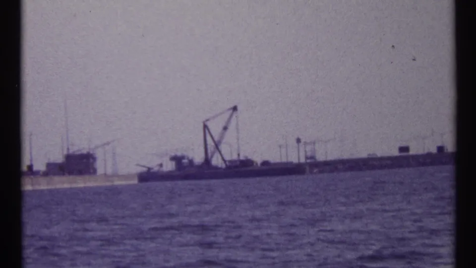 1967 Montreal harbor view with open water, a distant barge-like vessel, crane structure, lampposts, and low industrial buildings.