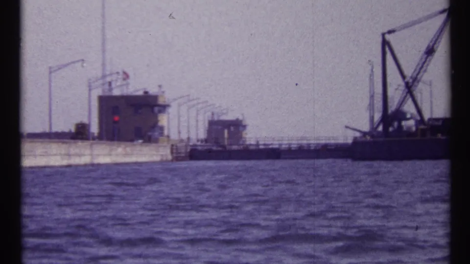 End frame of a 1967 Montreal waterway shot with lock buildings, guide walls, railings, and a crane barge under a pale sky.