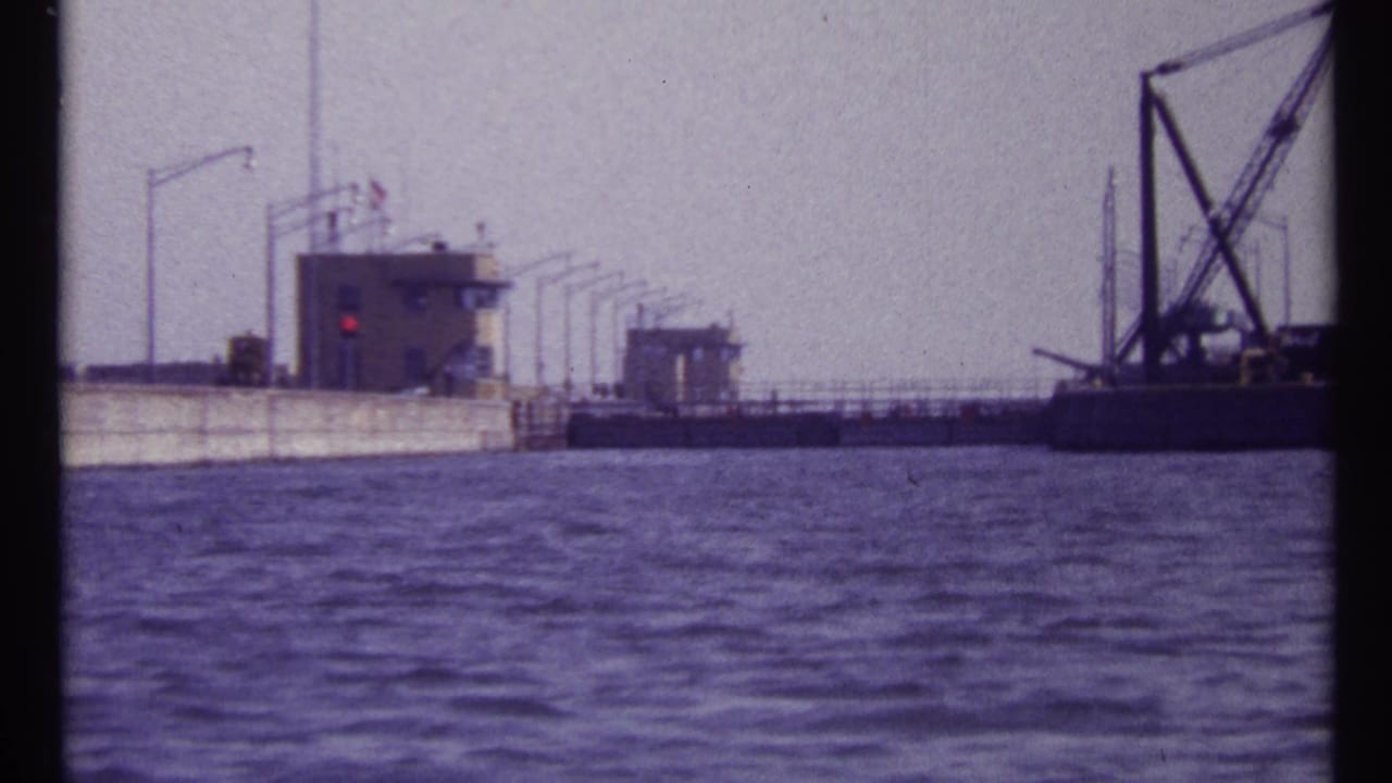 Concrete waterfront structure with a control building, red signal light, and crane barge across rippled water.
