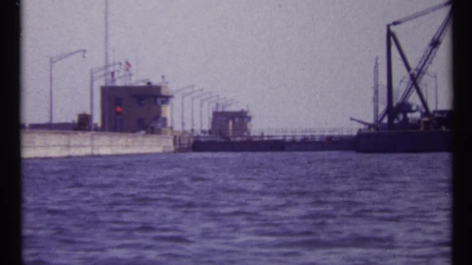 1967 Montreal home-movie frame showing a concrete lock or dam structure, small control building, red signal light, and crane barge across the water.