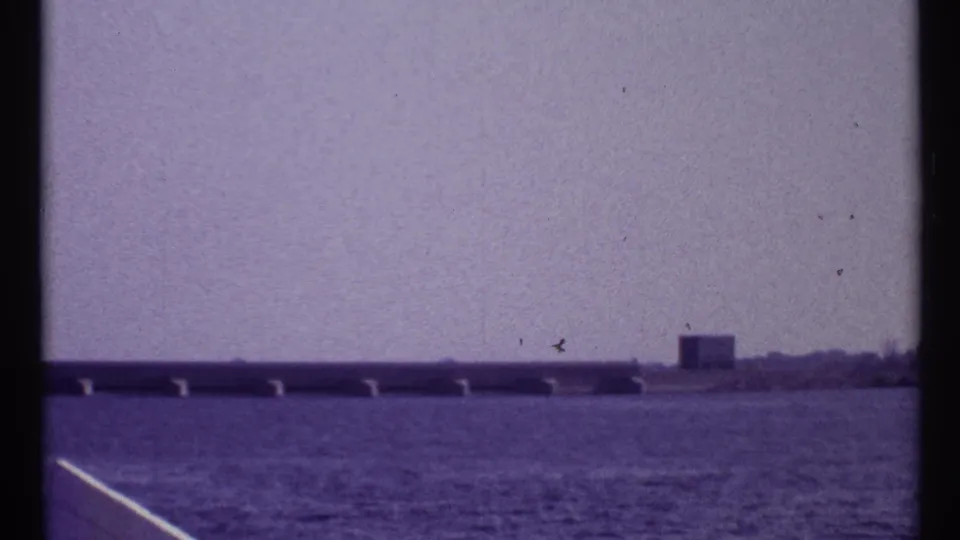 1967 Montreal home-movie frame showing a low bridge or causeway, open water, distant shoreline, and a small boxlike building.