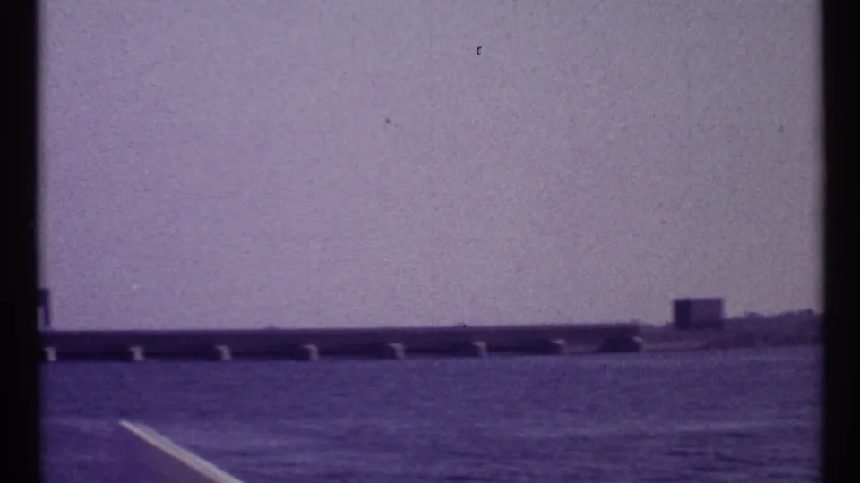 1967 Montreal waterway frame with rippled foreground water, a low bridge or causeway, and a small distant structure under a pale sky.