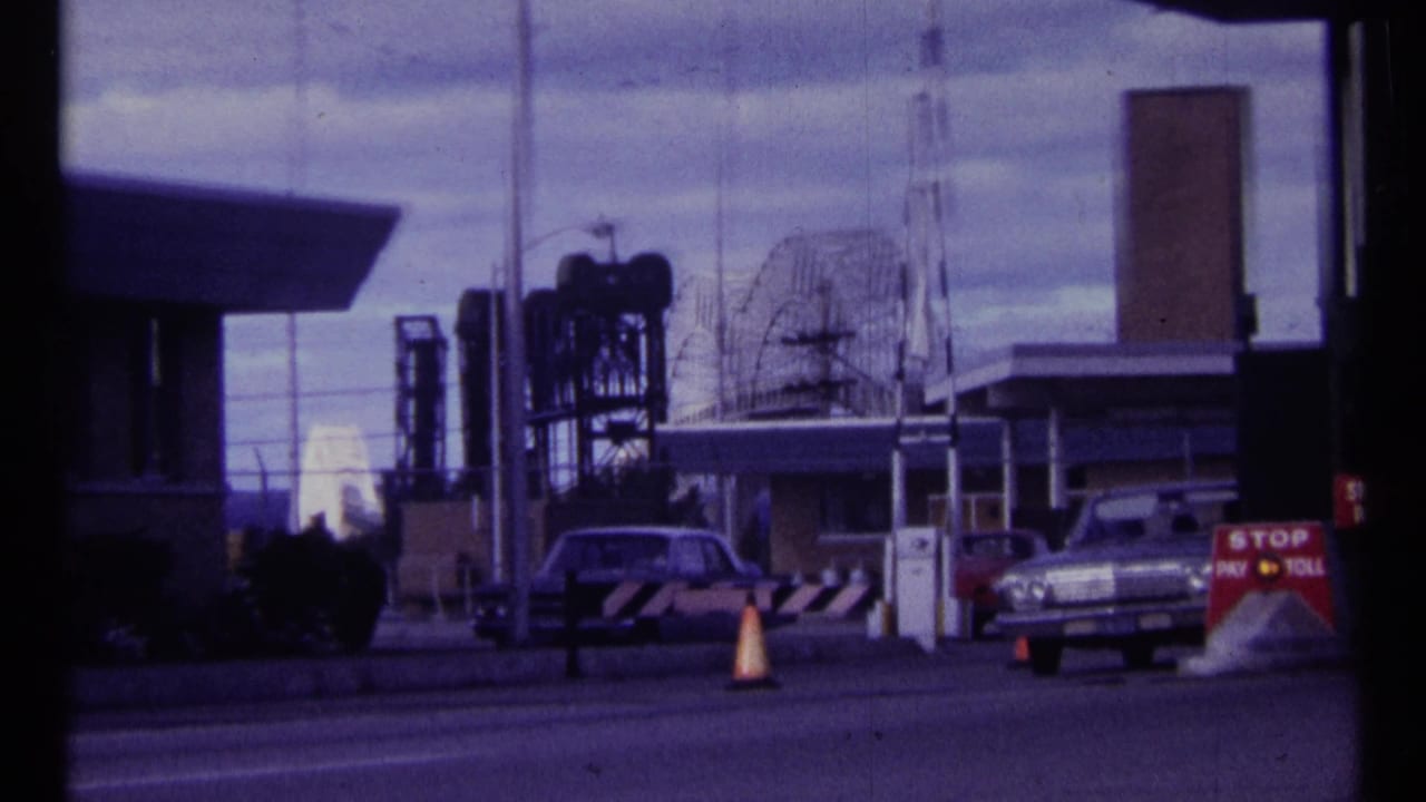 1967 Ottawa frame showing cars paused at a toll or security gate with cones, industrial structures, antennas, and a STOP PAY TOLL sign.