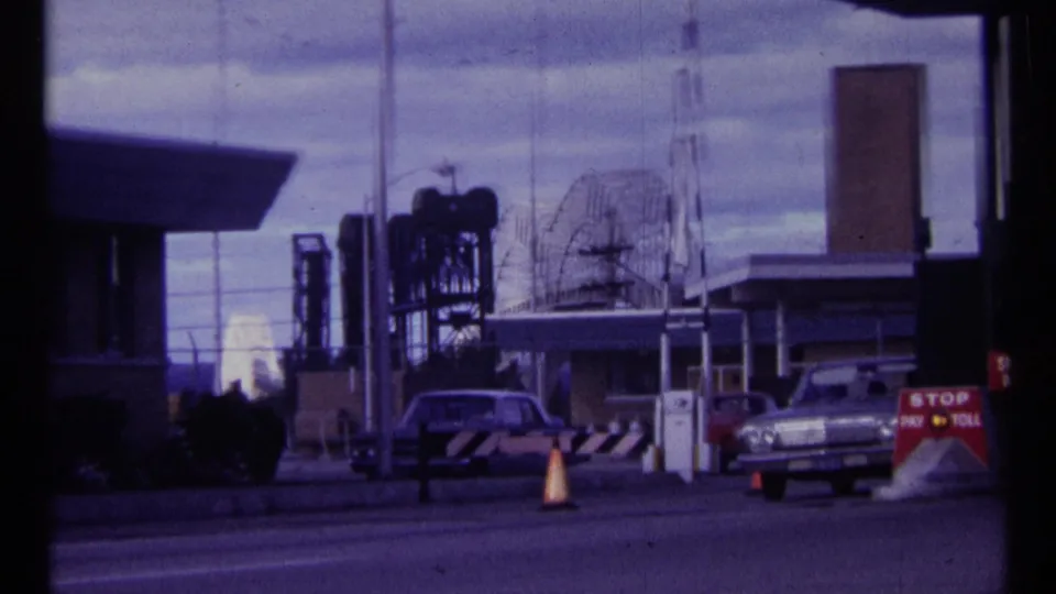 1967 Ottawa frame showing cars paused at a toll or security gate with cones, industrial structures, antennas, and a STOP PAY TOLL sign.