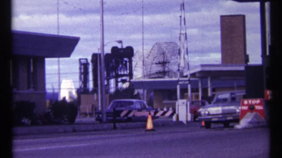 1967 Ottawa home-movie view of a car waiting near a toll gate, with cones, antennas, buildings, and industrial equipment visible.