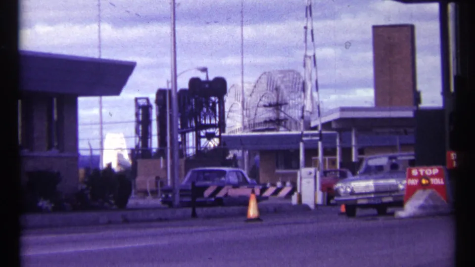 1967 Ottawa frame of cars at a toll or security checkpoint, with orange cones, a red sign, buildings, antennas, and industrial structures.