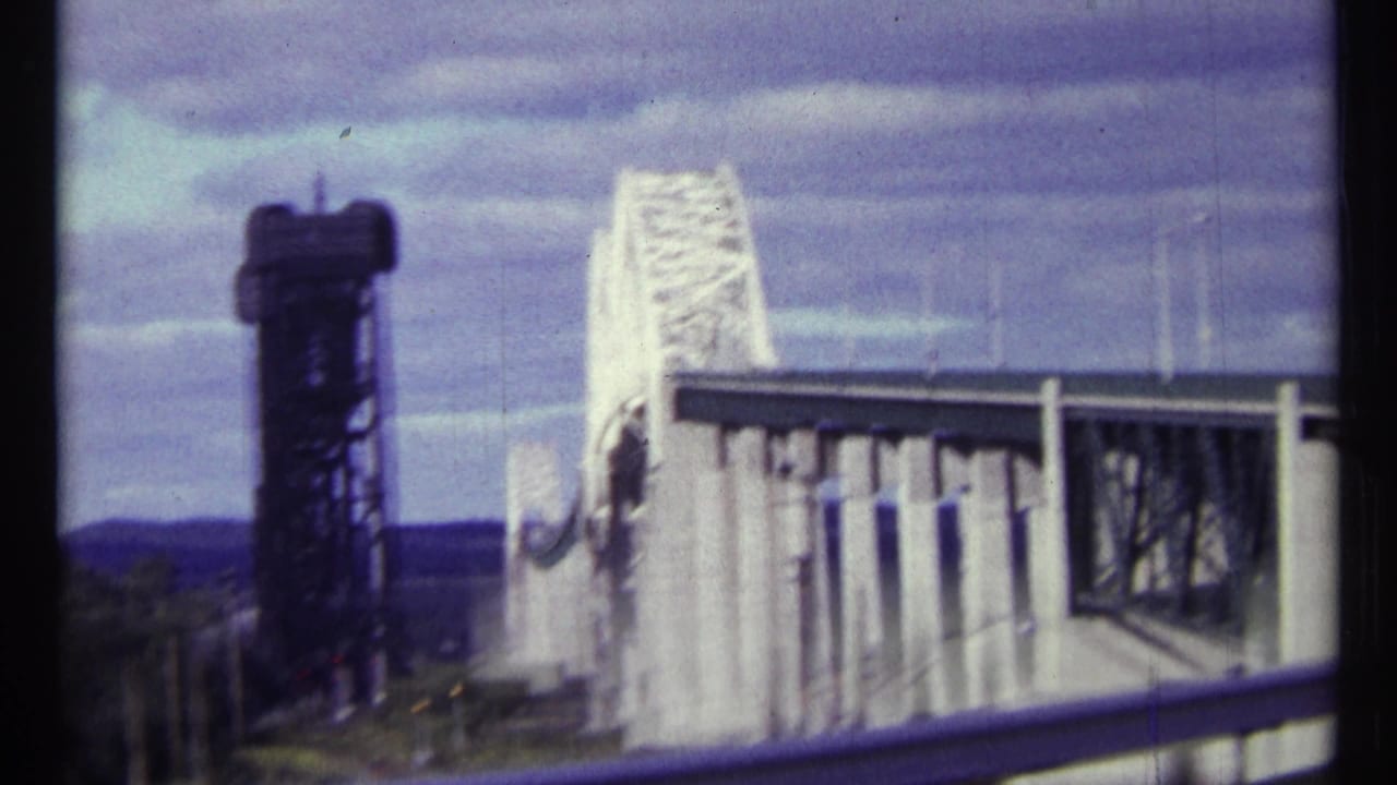 White truss bridge and dark control tower seen from a road with a guardrail under a cloudy sky.