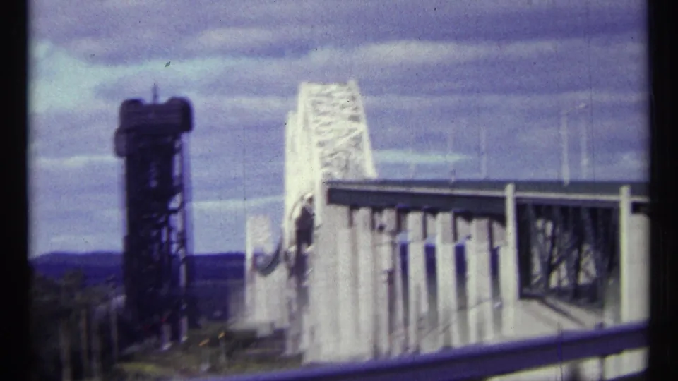 1967 Ottawa home-movie frame showing a white steel bridge, dark control tower, guardrail, and distant landscape from a moving road view.
