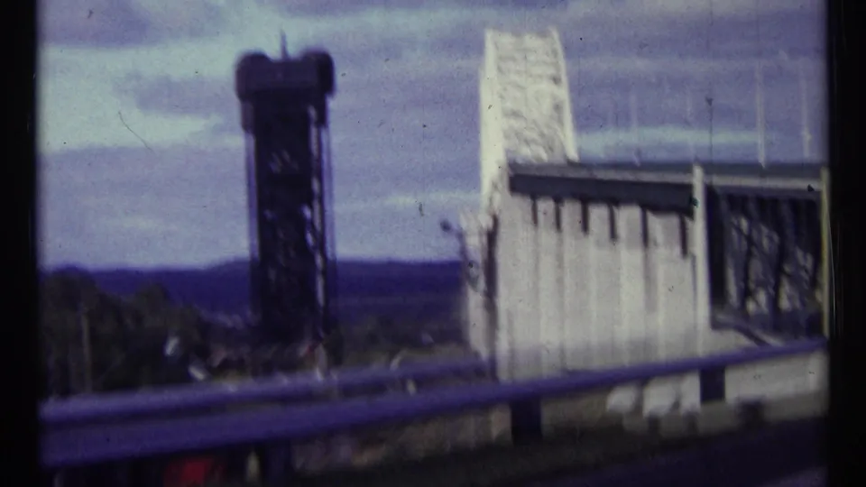 Close 1967 Ottawa road view of a dark bridge tower and white steel bridge, softened by motion blur and 8mm grain.
