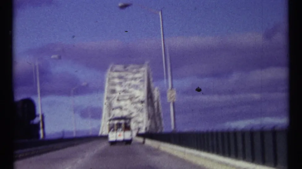 1967 Ottawa home-movie frame looking down the road toward a raised white bridge span with a small vehicle ahead under cloudy sky.