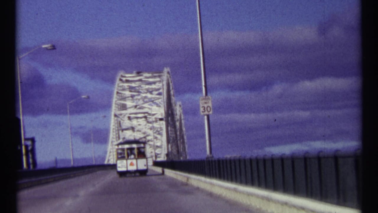 Roadway approaching a white truss bridge with a vehicle ahead and a speed sign at right.