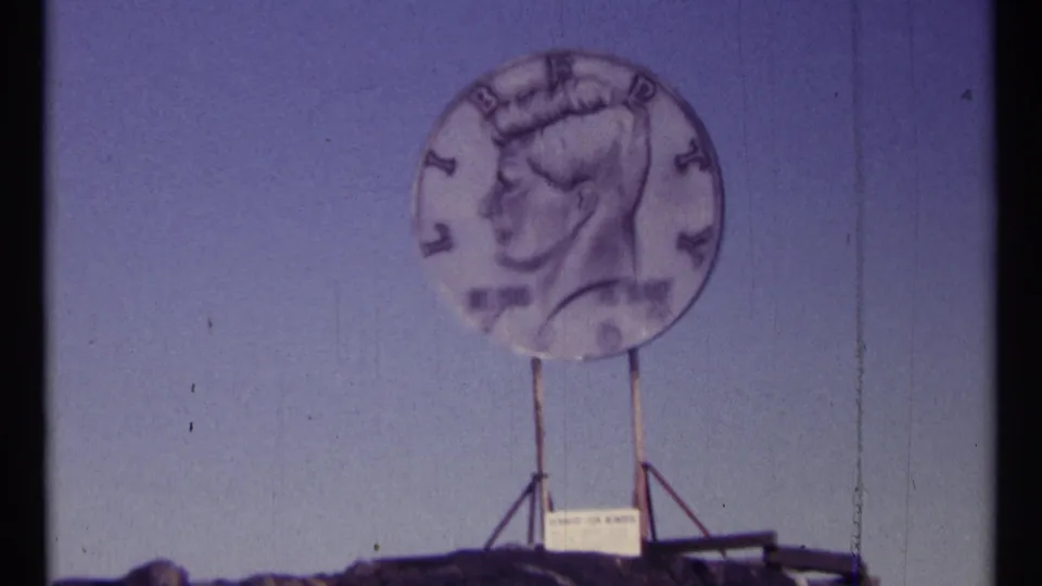 1967 Ottawa home-movie frame showing a large Kennedy half-dollar replica mounted above a roadside stand.