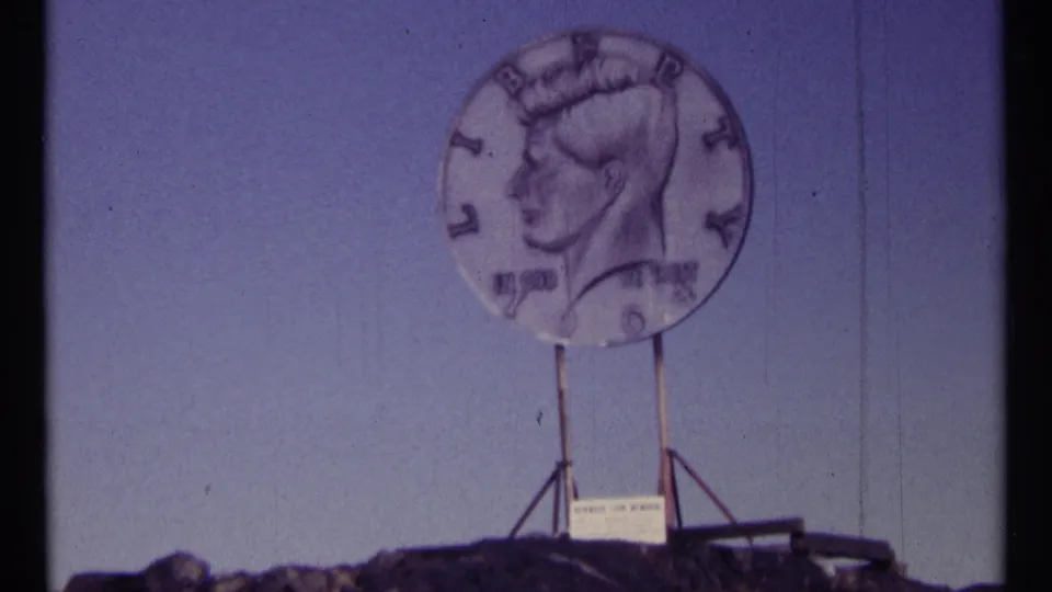 The giant Kennedy half-dollar replica is framed more centrally, with its support structure and small sign visible below.