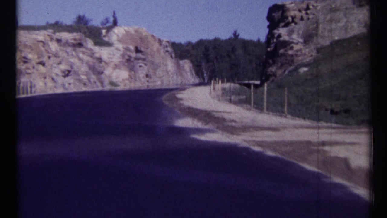 Curving asphalt road beside rock cliffs, forest, gravel shoulder, and roadside posts.