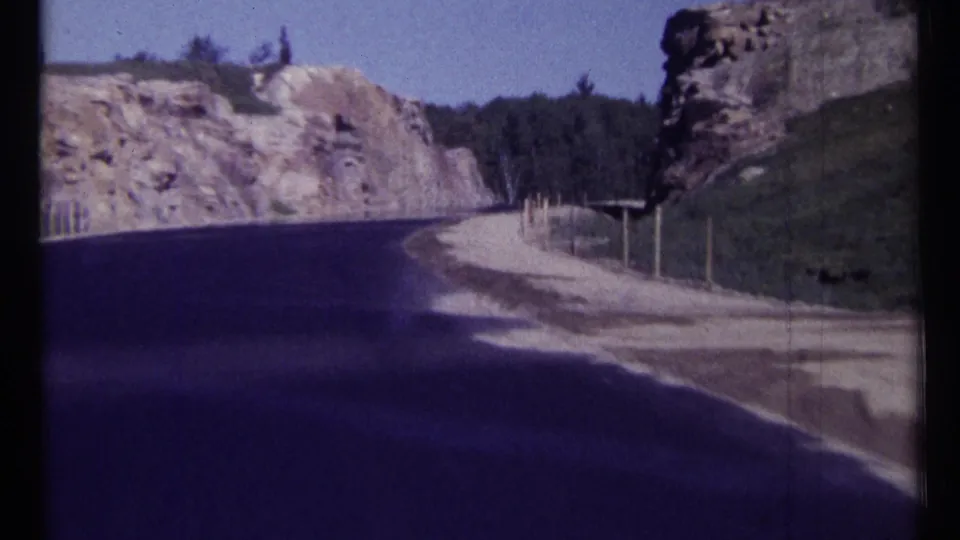 1967 Ottawa-area 8mm frame showing a winding road between rocky cliffs and trees, with a gravel shoulder and simple roadside posts.