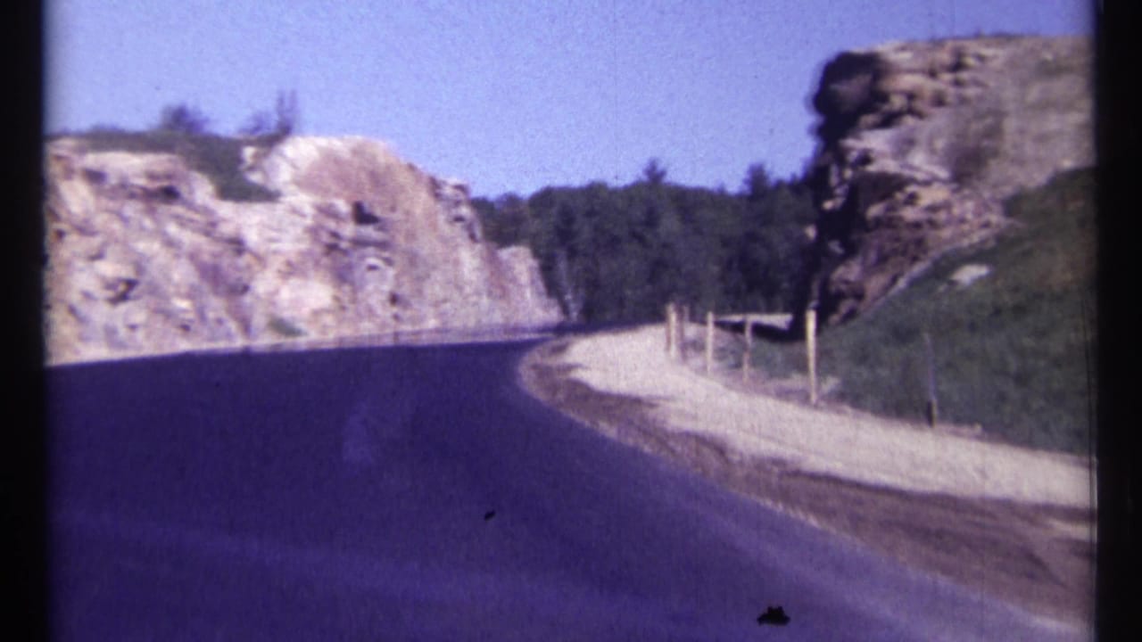 Road bends around pale rock cuts with trees and a fenced gravel shoulder.