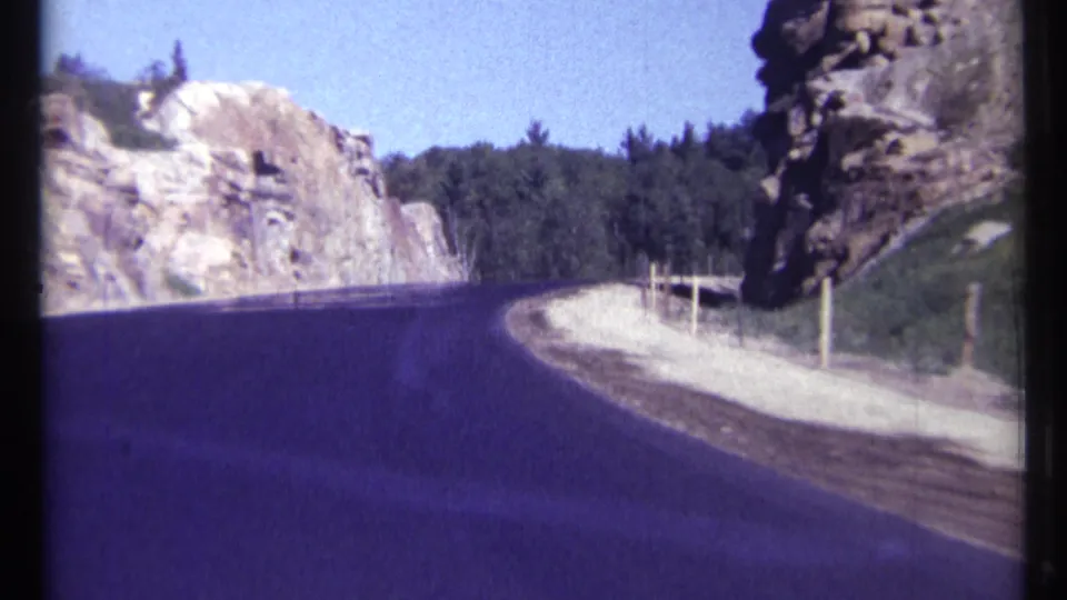 1967 Ottawa-area 8mm frame with the road filling the foreground as it approaches rock cliffs, a tree line, and a posted gravel shoulder.