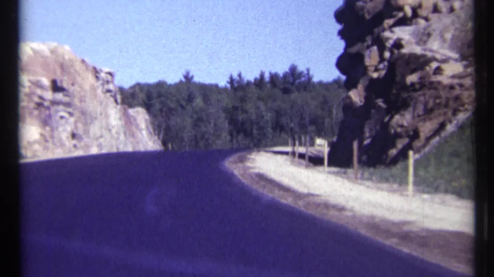 1967 Ottawa-area home-movie frame showing a curving road through a rocky forested cut, with roadside posts and a small sign shape in the distance.