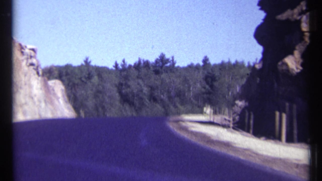 Curving road with rock face, trees, posts, and blue sky.