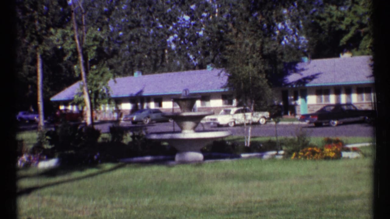 Motel grounds with a central fountain, lawn, trees, parked cars, and single-story rooms.