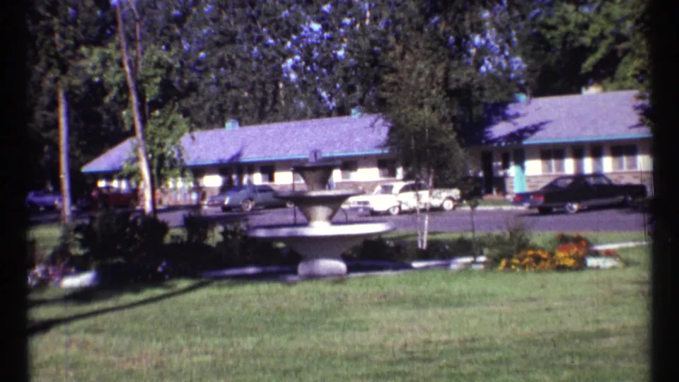 1967 motel exterior showing a fountain in the foreground, lawn and flower beds, and cars parked outside single-story rooms.