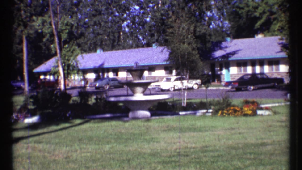 Motel grounds with a fountain, lawns, flower beds, parked cars, and a one-story building.