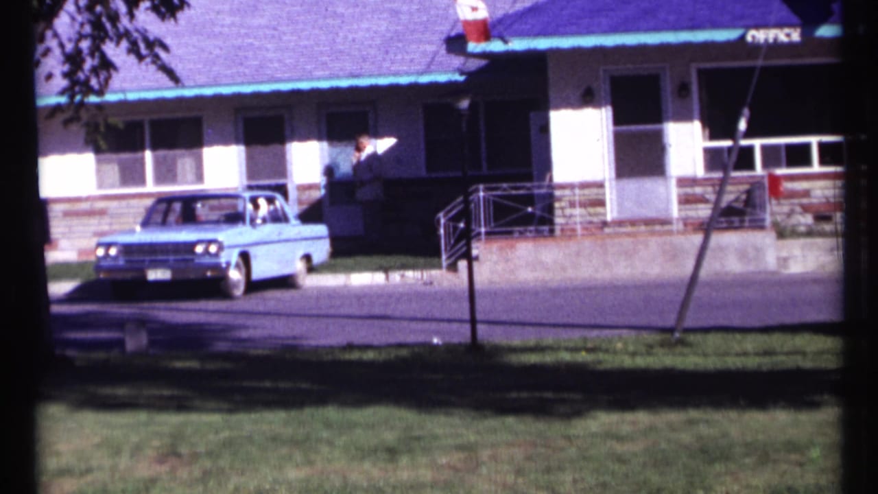 1967 Ottawa frame of a light blue vintage sedan passing a motel office entrance while a person stands near the doorway.