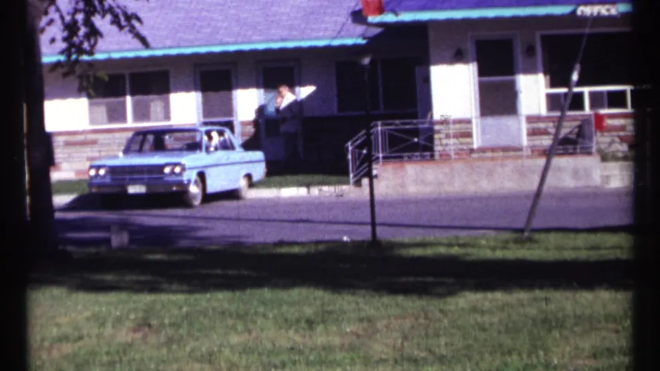 1967 Ottawa frame with a blue vintage sedan outside a motel office, a person by the doorway, and grass in the foreground.