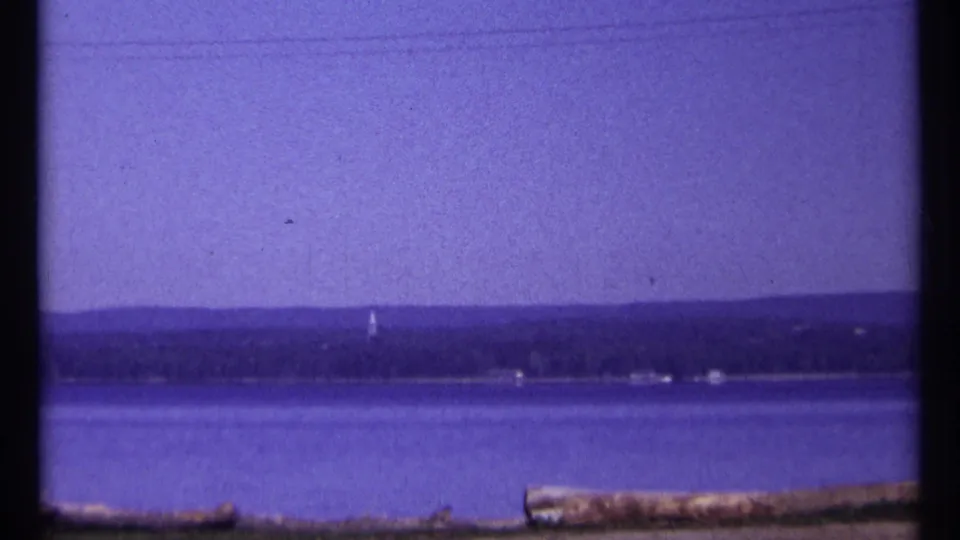 1967 Ottawa-area 8mm frame with a broad water view, distant trees and small structures, overhead wires, and logs along the foreground edge.