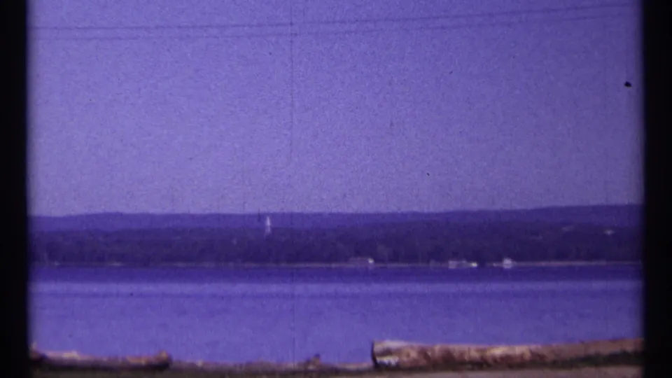 Ottawa-area 1967 home-movie view across calm water toward a low wooded horizon, with faint buildings and logs in the foreground.