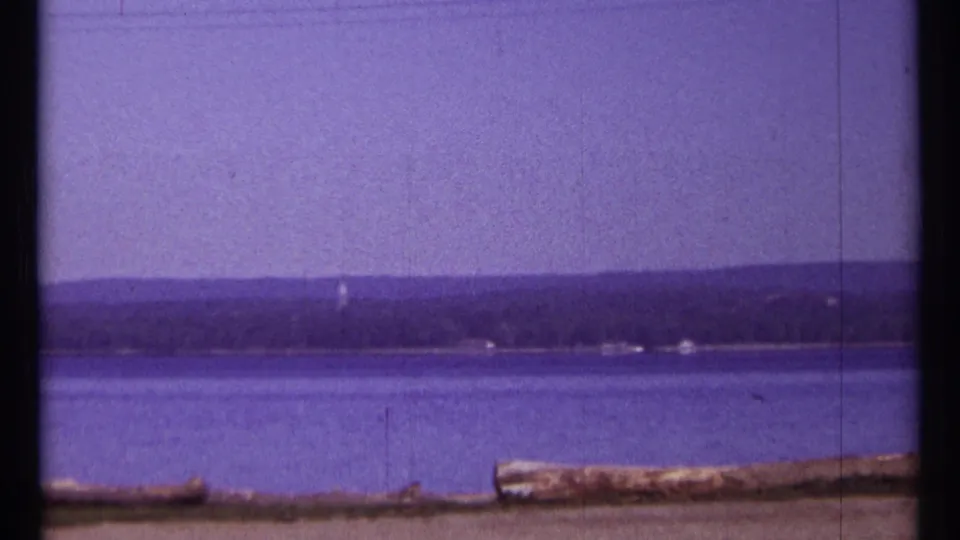 1967 Ottawa-area waterfront frame with blue water, a dark distant shoreline, a small pale vertical detail, and logs at the bottom edge.