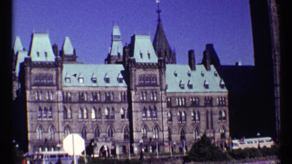 1967 Ottawa Parliament exterior from a visitor viewpoint, with arched windows, pale rooflines, small people, and a sign in the foreground.