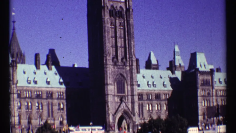 1967 Ottawa home-movie frame showing the Peace Tower and Parliament Hill stone buildings with pale roofs and blue sky.