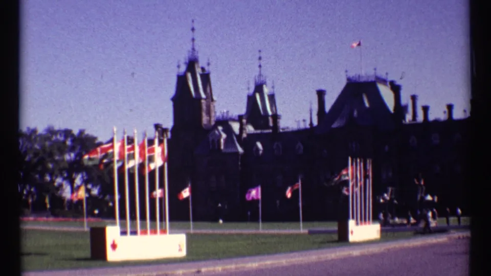 1967 Ottawa Parliament Hill view with Canadian and provincial flags waving before dark stone buildings.