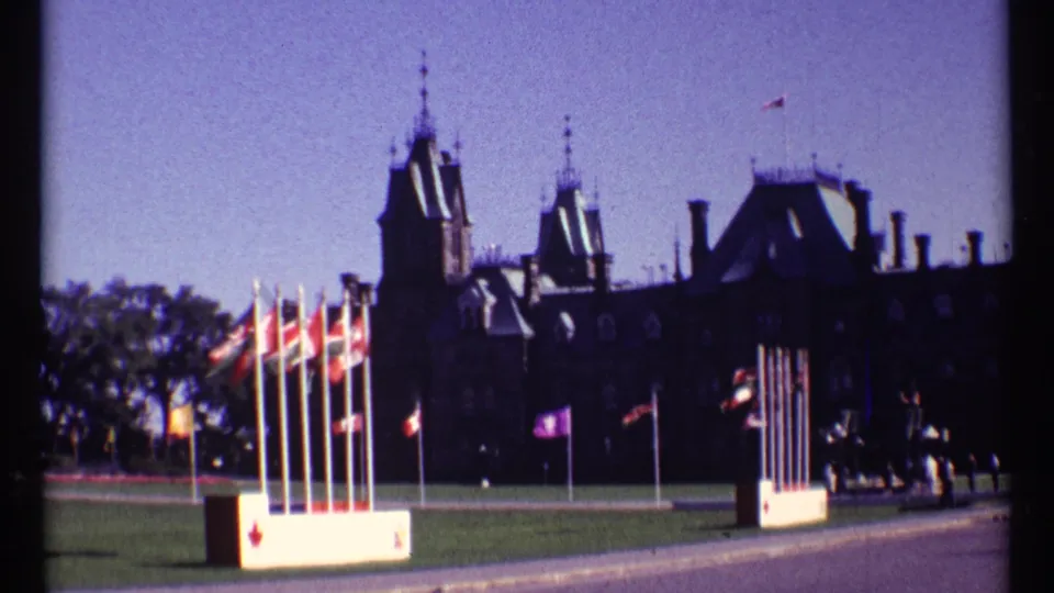 1967 8mm view of Ottawa Parliament buildings with multiple flags in the foreground under a clear sky.