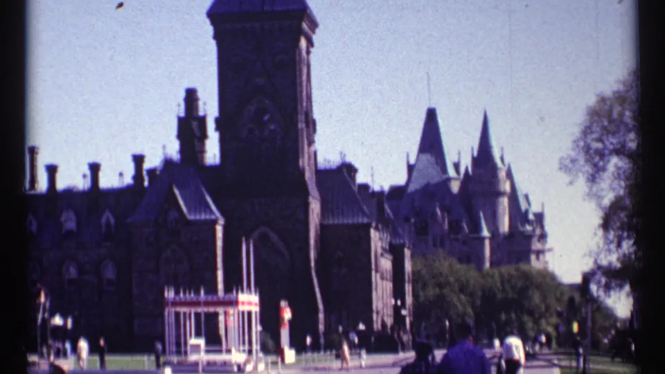 Visitor-shot 8mm frame of Ottawa Parliament architecture, pointed rooftops, trees, and people walking near the grounds.