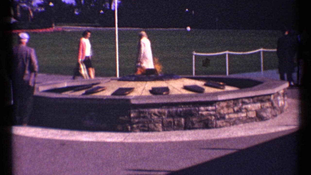 1967 Ottawa home-movie frame showing suited men and women in dresses around a circular stone flame feature with grass and railings behind them.