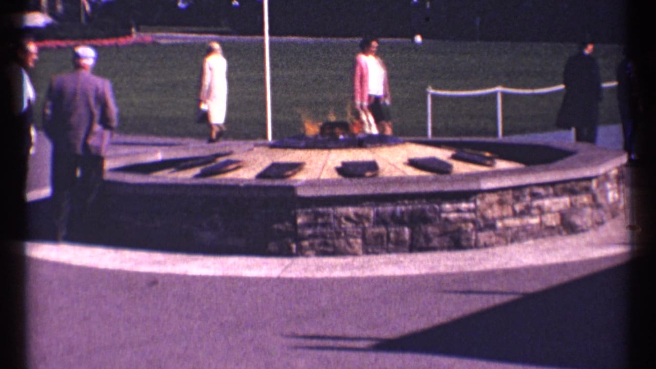 People in formal street clothes stand and walk around the flame monument beside a lawn.