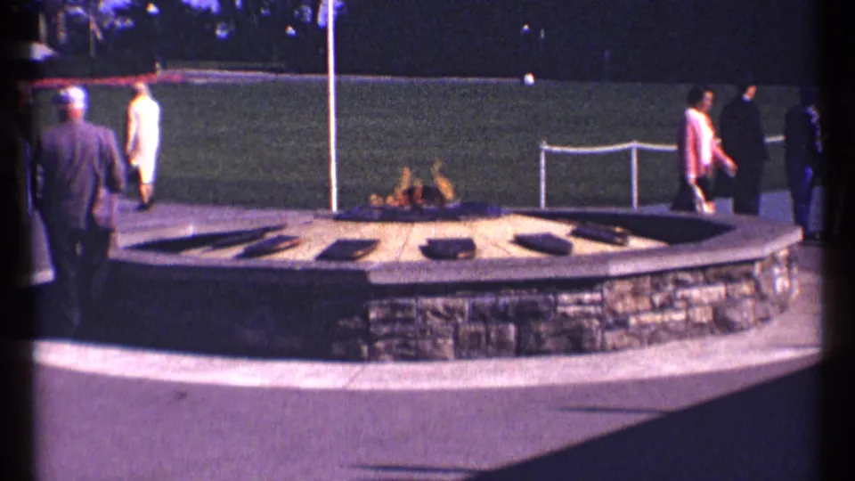 Home-movie view of the Ottawa public flame area, with visitors in 1960s clothing moving past the stone feature and trimmed grass.