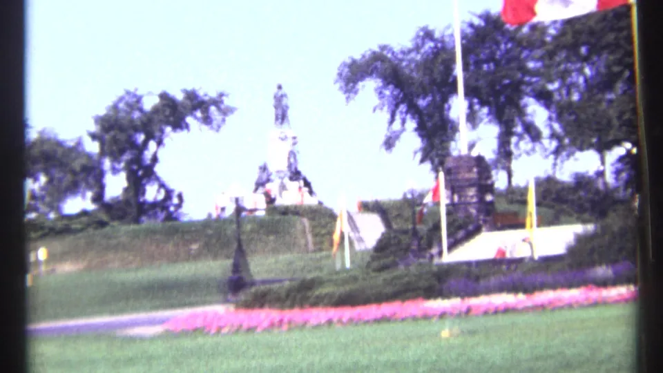 Wide 1967 Ottawa park view with a distant monument, flagpoles, trees, lawn, and a pink flower bed under bright daylight.
