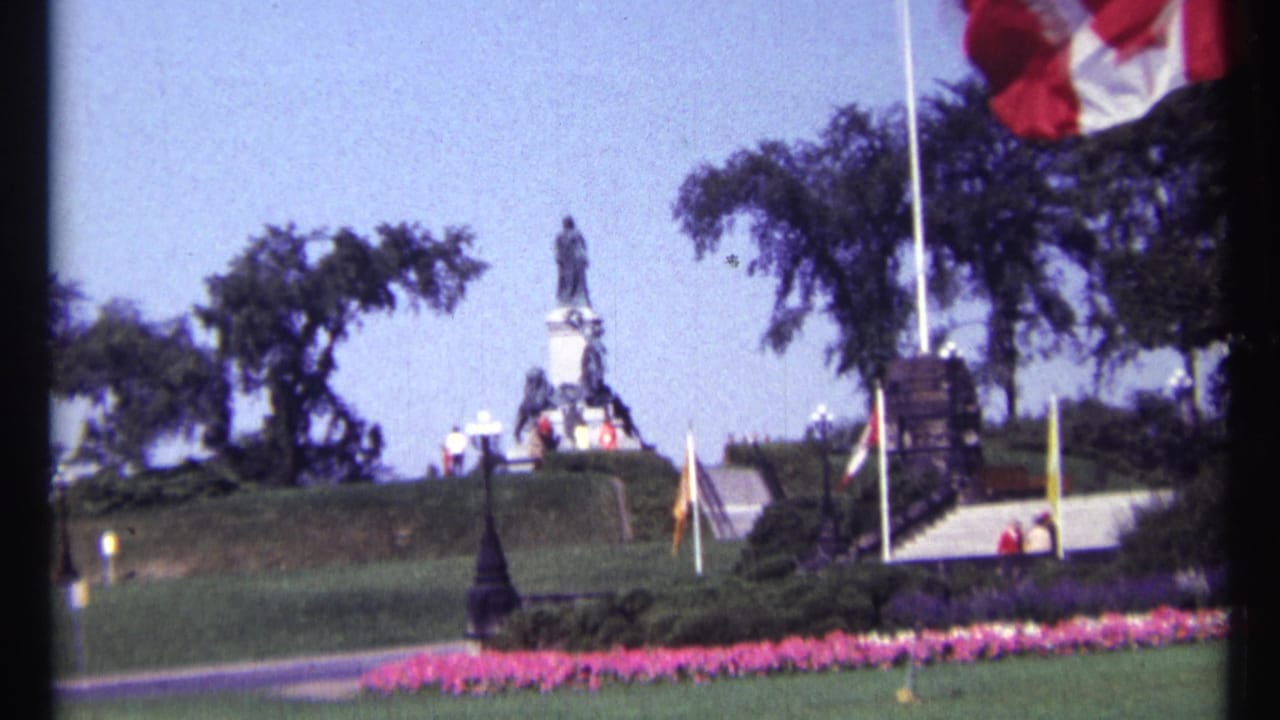 Canadian flag above a landscaped public monument with lawns, paths, trees and flowerbeds.