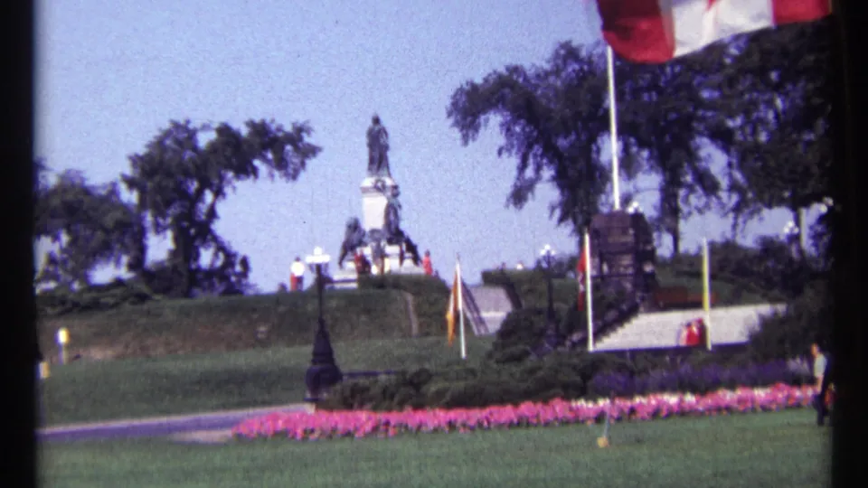 1967 Ottawa home-movie frame of formal grounds around a monument, with a Canadian flag, stairway, trees and small distant figures.