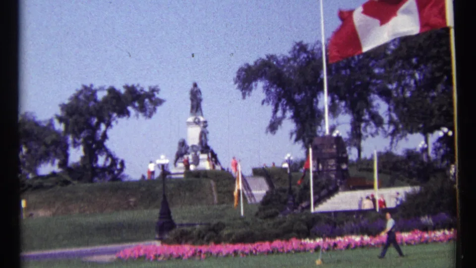 1967 Ottawa 8mm frame showing a person crossing landscaped grounds below a monument with flagpoles, trees and bright flowerbeds.