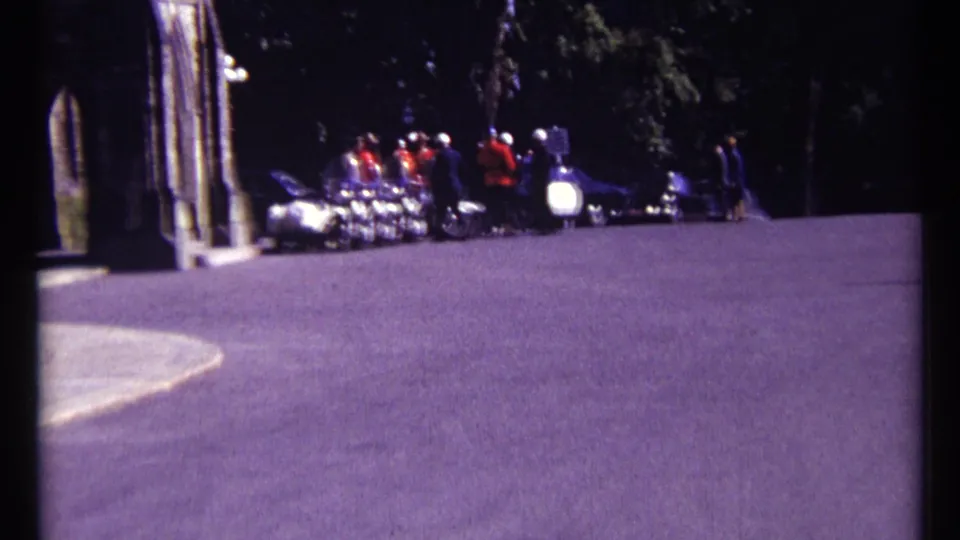 1967 Ottawa home-movie frame showing a uniformed group, possibly musicians, near parked vehicles by a paved drive and stone structure.